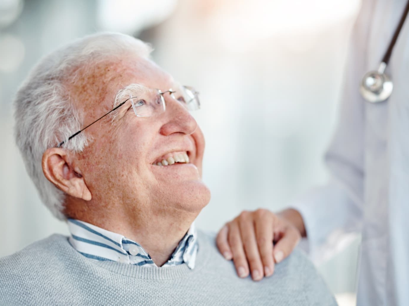 An older patient in a grey sweater smiling up at a clinician in a white coat whose hand rests on his shoulder